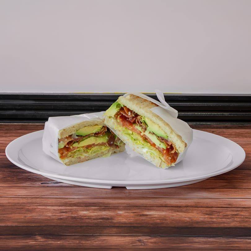 Two halves of a sandwich with bacon, avocado, lettuce, and tomato on a white plate.